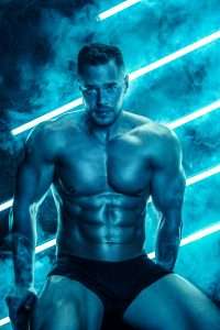 male strip shows Manhattan: Shot of muscular guy with perfect body sitting against background with neon lighting.